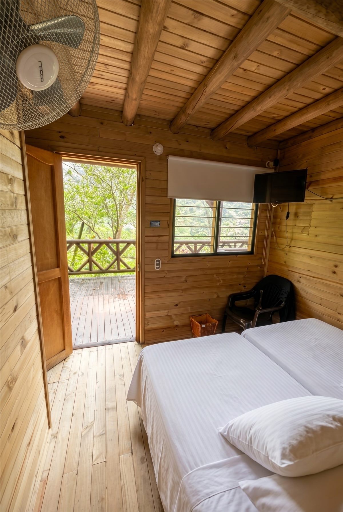 Habitación con aire de día: paredes y techo en madera, camas con sábanas blancas, televisor, ventilador y puerta al balcón compartido